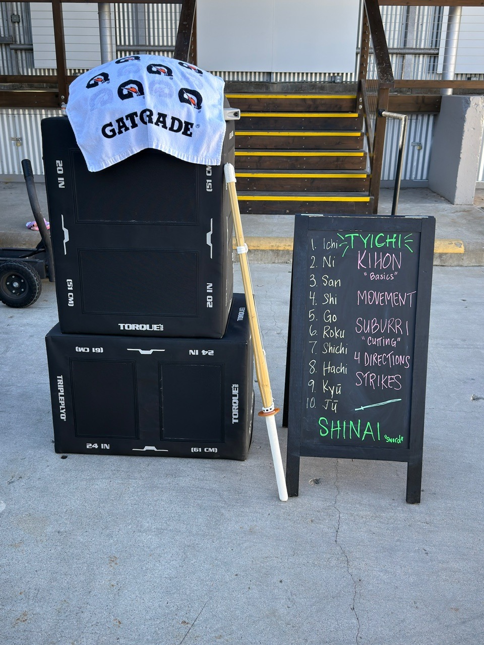 A-frame chalkboard at Lilley Fitness reading TYICHI: Kihon Basics, Movement, Suburri Cutting, 4 Directions Strikes, Shinai Sword, and counts 1 to 10 in Japanese, beside a bamboo shinai leaning against plyo boxes.